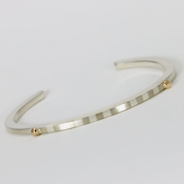 Cuff bracelet fabricated in sterling silver and has two 14k gold balls with a striped pattern between them.