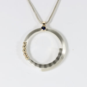 Spiral circular Pendant with a striped pattern and a sapphire. Fabricated in sterling silver with solid 14k gold balls