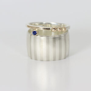 Wide sterling silver ring with a subtle striped pattern. There is 14k gold and a sapphire.