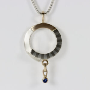 Mixed metal pendant with a dangling sapphire. Hand fabricated in sterling silver and 14k gold. Marriage of metals subtle striped pattern detail.