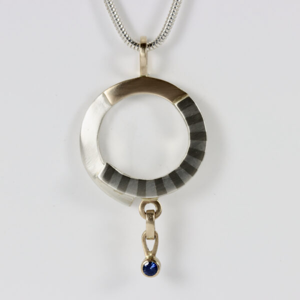 Mixed metal pendant with a dangling sapphire. Hand fabricated in sterling silver and 14k gold. Marriage of metals subtle striped pattern detail.