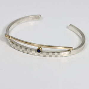 A cuff bracelet hand-fabricated in sterling silver and 14k gold. Details include a checkerboard pattern and a Sapphire.