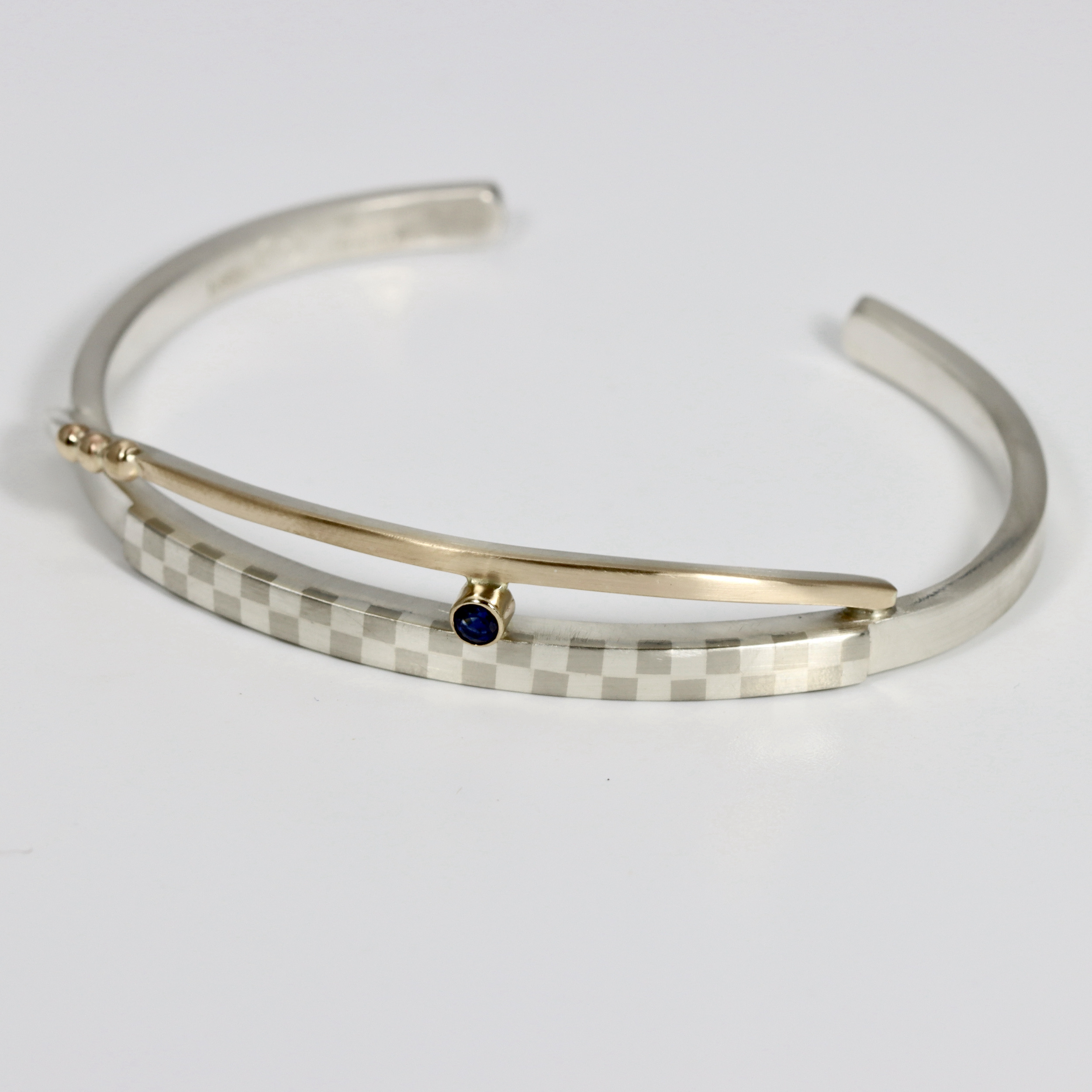 A cuff bracelet hand-fabricated in sterling silver and 14k gold. Details include a checkerboard pattern and a Sapphire.