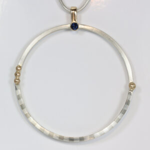 Mixed-metal circular pendant with a striped pattern and a sapphire. Hand-fabricated in sterling silver and 14k gold.