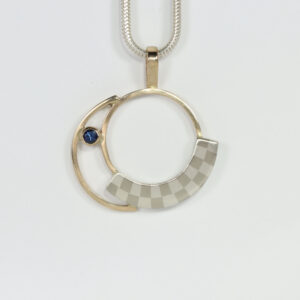A circular pendant with a sapphire. Hand-fabricated in sterling silver and 14k gold. A subtle checkerboard pattern contrast lovely with the 14k yellow gold and blue sapphire.