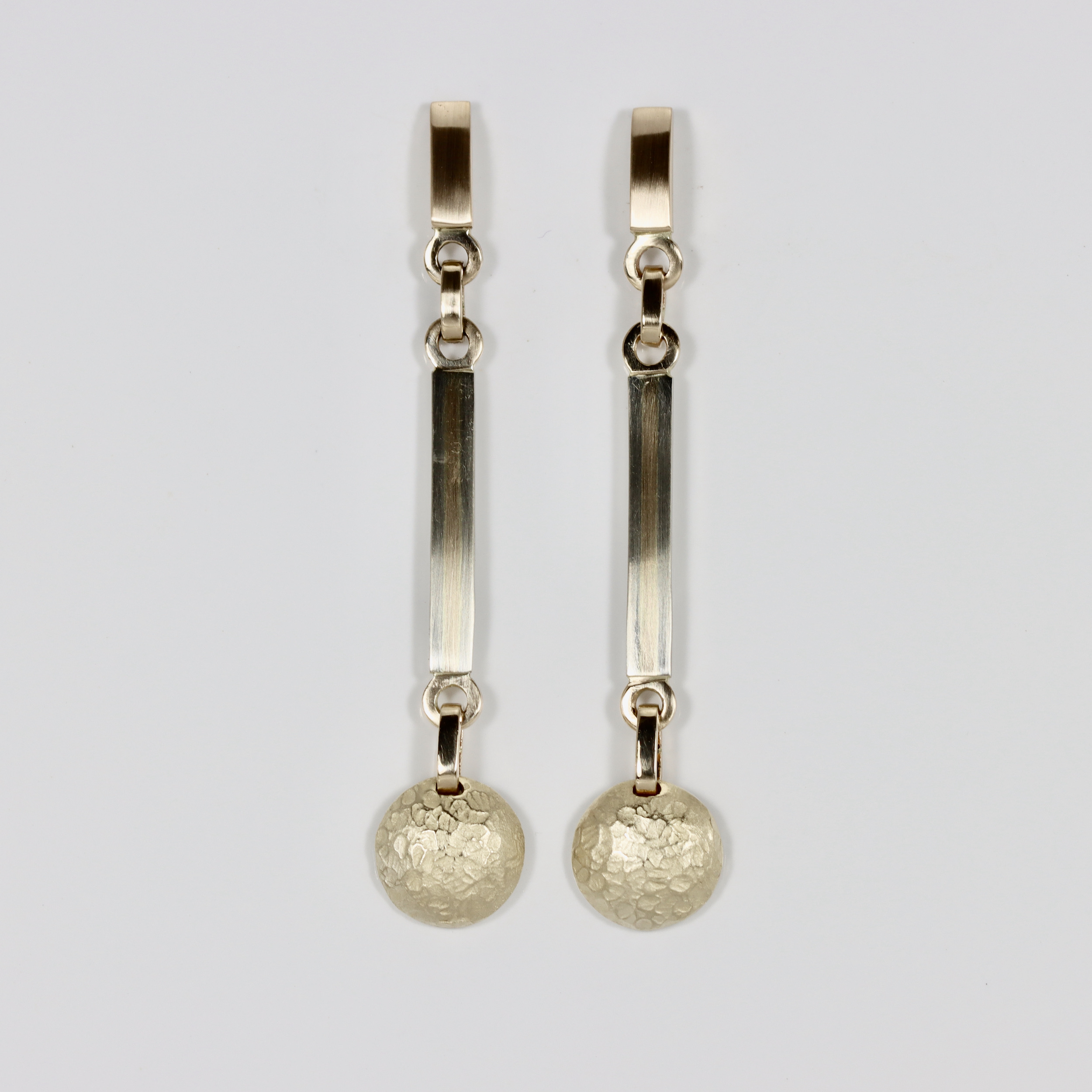 A pair of long, multi-tiered 14k gold dangles. The earrings feature a top post that secures to the ear, holding a middle section made of a long, narrow drop component with an alternating pattern of 14k yellow gold and white gold stripes. Suspended from the bottom of this slender bar is a domed circle with a textured finish.
