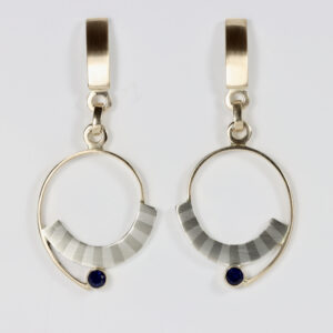 A pair of handcrafted post-and-dangle earrings showcasing mixed-metal artistry. The top consists of a simple post stud. Hanging below is a flat, circular geometric drop featuring a seamless "marriage of metals" design with alternating sterling silver stripes. A delicate 14k gold wire spiral around the circular disk, and a single, vibrant deep blue sapphire is meticulously bezel-set at the bottom center of the dangling piece.