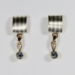 A single post-and-dangle earring featuring a striped, domed rectangular post. The metal post is detailed with fine alternating stripes. Suspended from the bottom of the rectangle is a single, faceted AAA-round-shaped blue-grey spinel gemstone that hangs freely, catching the light.